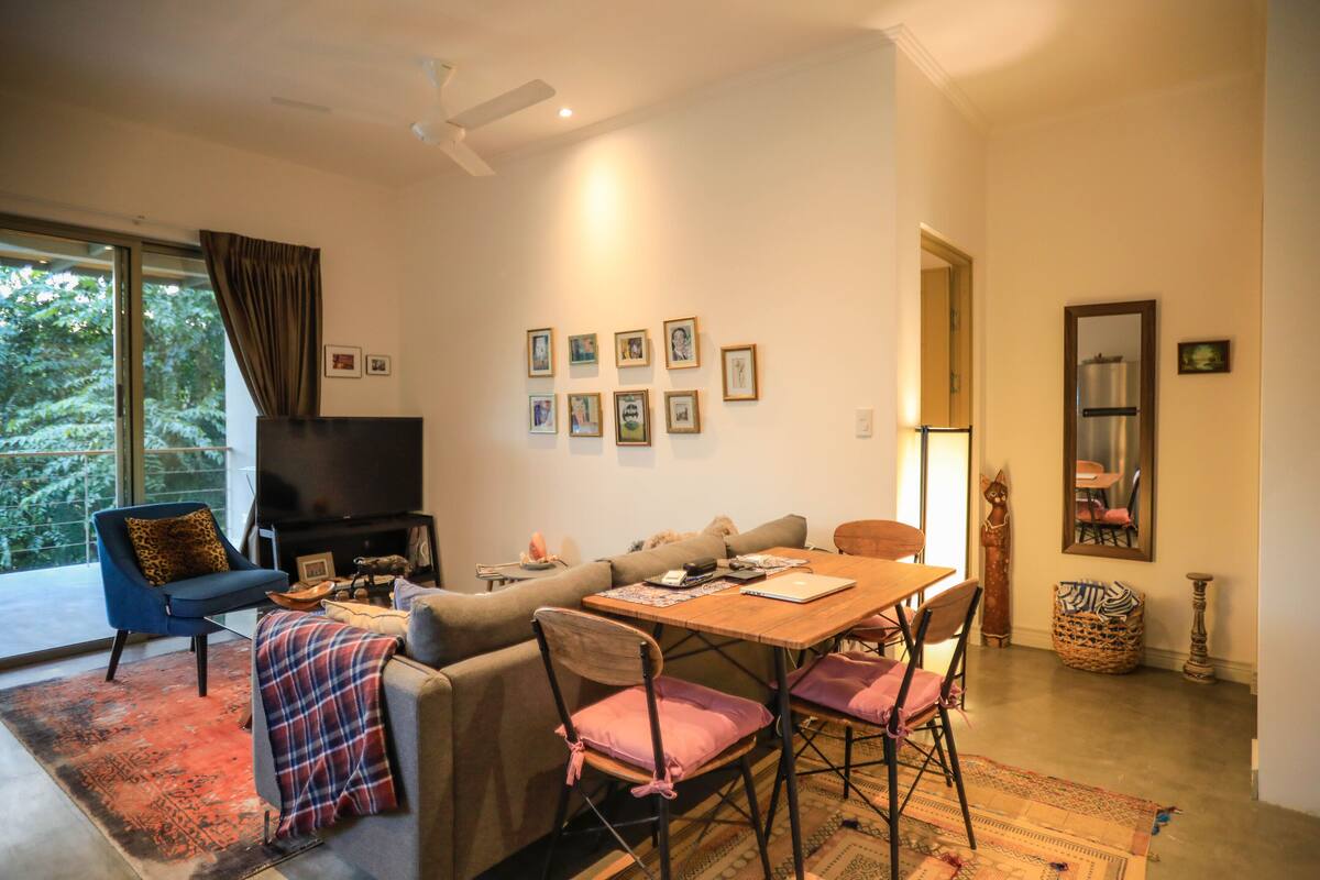 Uma sala de estar com sofá, mesa e cadeiras. Há uma televisão e uma janela com cortinas.