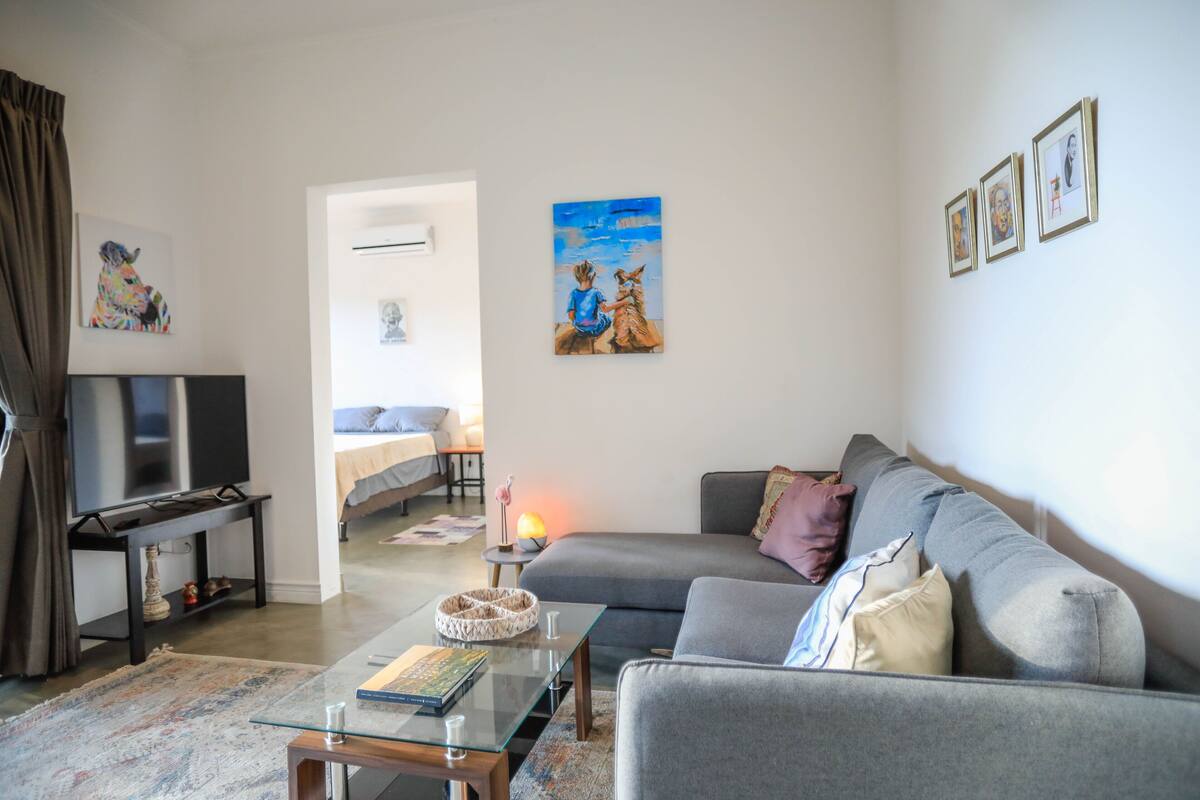 Uma sala de estar com um sofá cinzento, uma mesa de centro de vidro, uma TV sobre um suporte e obras de arte nas paredes.
