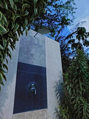 A modern outdoor water feature with a metal spout, surrounded by lush greenery and a clear blue sky.