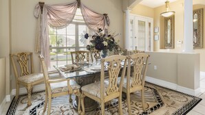 A dining area with a glass table surrounded by ornate chairs, a floral centerpiece, and a view of the outdoors through large windows with draped curtains. - Closest to Disney by VillaDirect (Kissimmee)