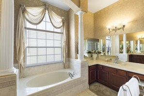 A bathroom with a large bathtub, a vanity with a mirror, and a window with curtains. - Closest to Disney by VillaDirect (Kissimmee)