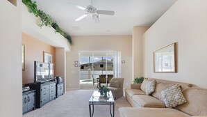 A living room with a beige sofa, a coffee table, a TV, and a ceiling fan. There is a sliding glass door leading to a patio area.