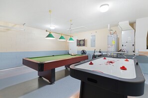 A pool table with a foosball table, a dartboard, and a fan in a room with a brick wall and a white ceiling.