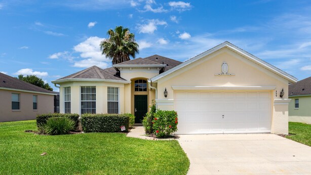 A single-story house with a garage, a front door, and a small front yard with a palm tree and bushes.