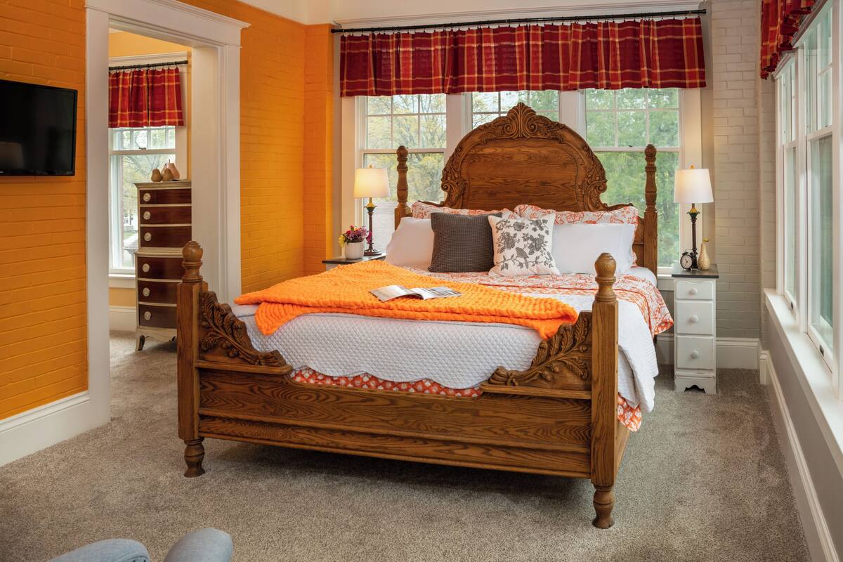 Un dormitorio con una cama de madera, dos mesitas de noche con lámparas, un aparador y una ventana con un adorno a cuadros.