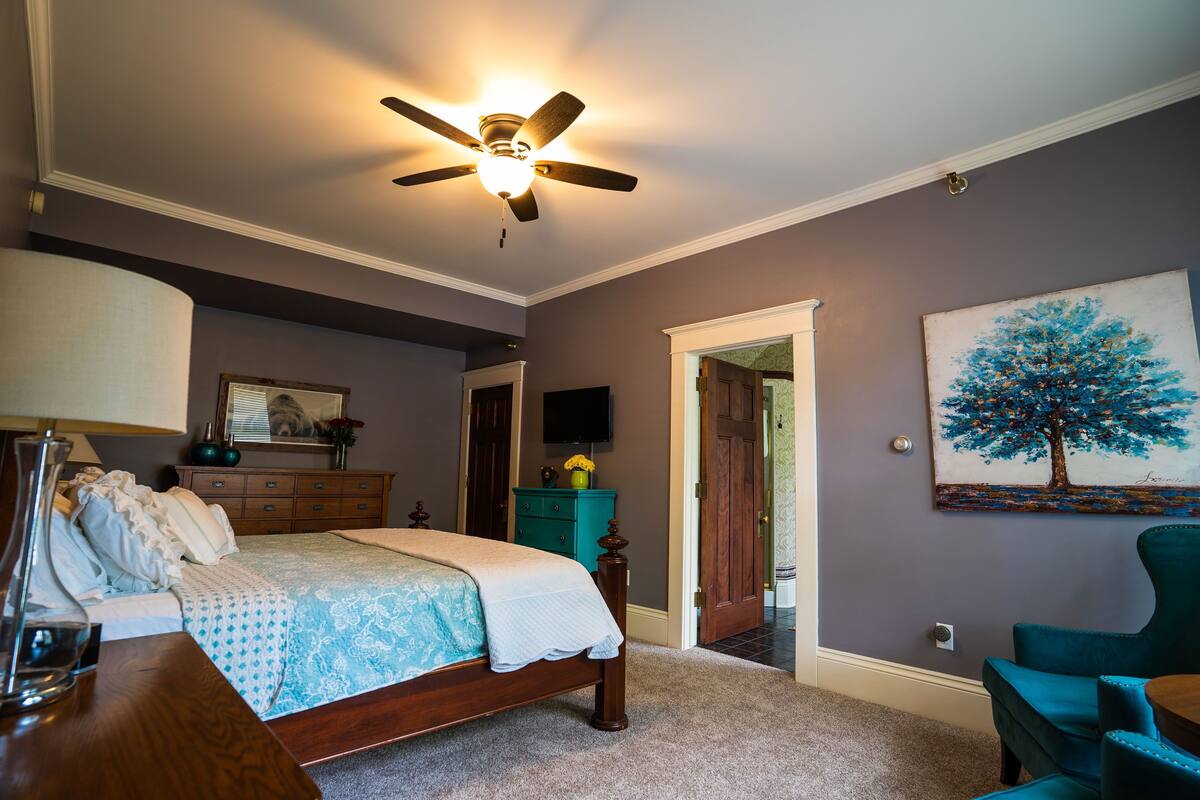 Un dormitorio con cama, ventilador de techo, cómoda, silla y un cuadro en la pared.