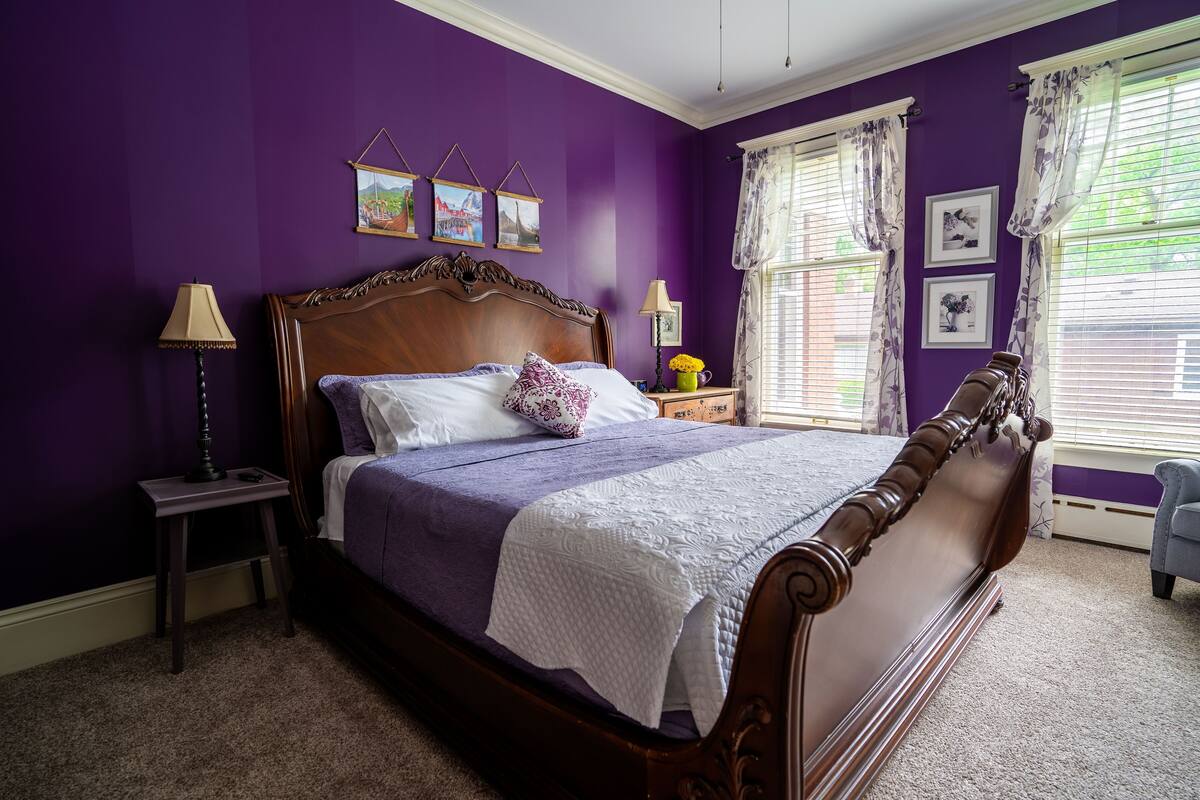 Un dormitorio con una cama grande, paredes moradas y una ventana con cortinas.