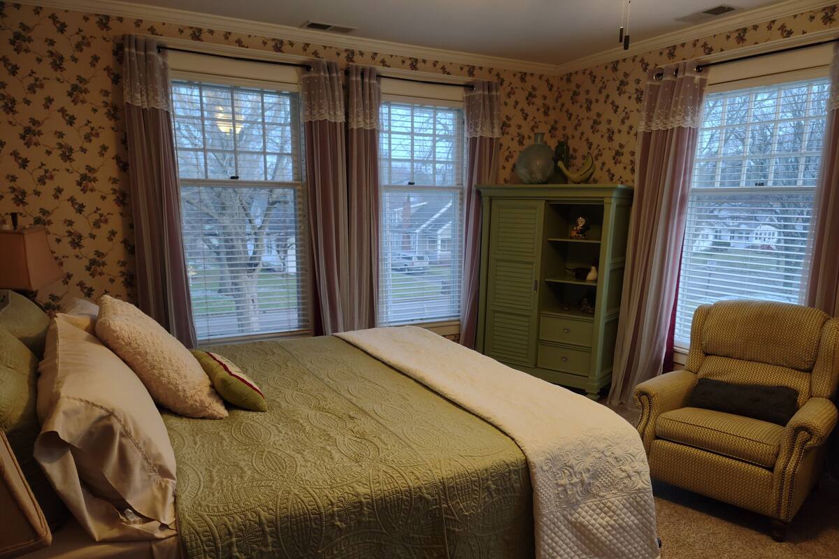 Un dormitorio con cama, armario verde, sillón amarillo y papel tapiz floral.