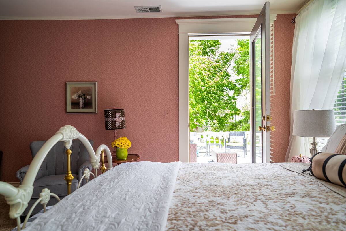 Un dormitorio con cama, una silla, un cuadro en la pared y una ventana con vista a árboles.