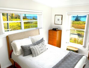 A bedroom with a bed, a dresser, and two windows with scenic views.