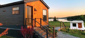 A cabin with a wooden exterior, a number '663' on the door, a small porch with a railing, and a view of a lake and forest in the background. - Lovely ocean views, hiking & lobster boat lounge (Machiasport)