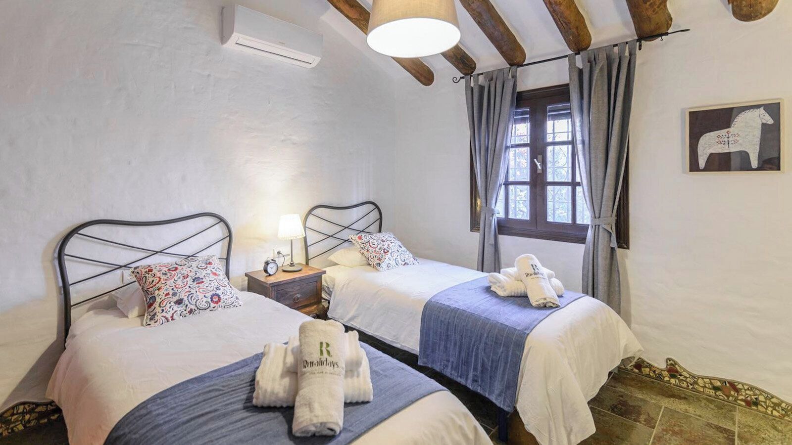 A bedroom with two beds, a nightstand with a lamp, a window with curtains, and a wall-mounted air conditioner.