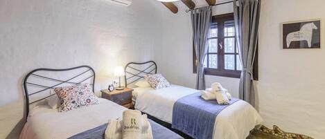 A bedroom with two beds, a nightstand with a lamp, a window with curtains, and a wall-mounted air conditioner.