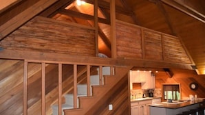 Uma escada de madeira leva a um andar superior, com uma área de cozinha e banquetas de bar, e um relógio na parede.