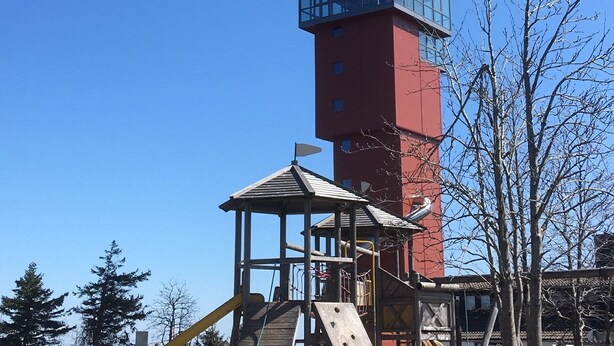 Un parque infantil con tobogán y estructuras para escalar, una torre roja con sección de vidrio y árboles sin hojas.
