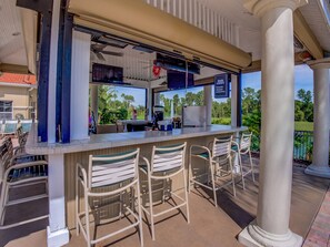 Uma área de bar ao ar livre, com balcão, banquetas e vista para o verde ao redor.