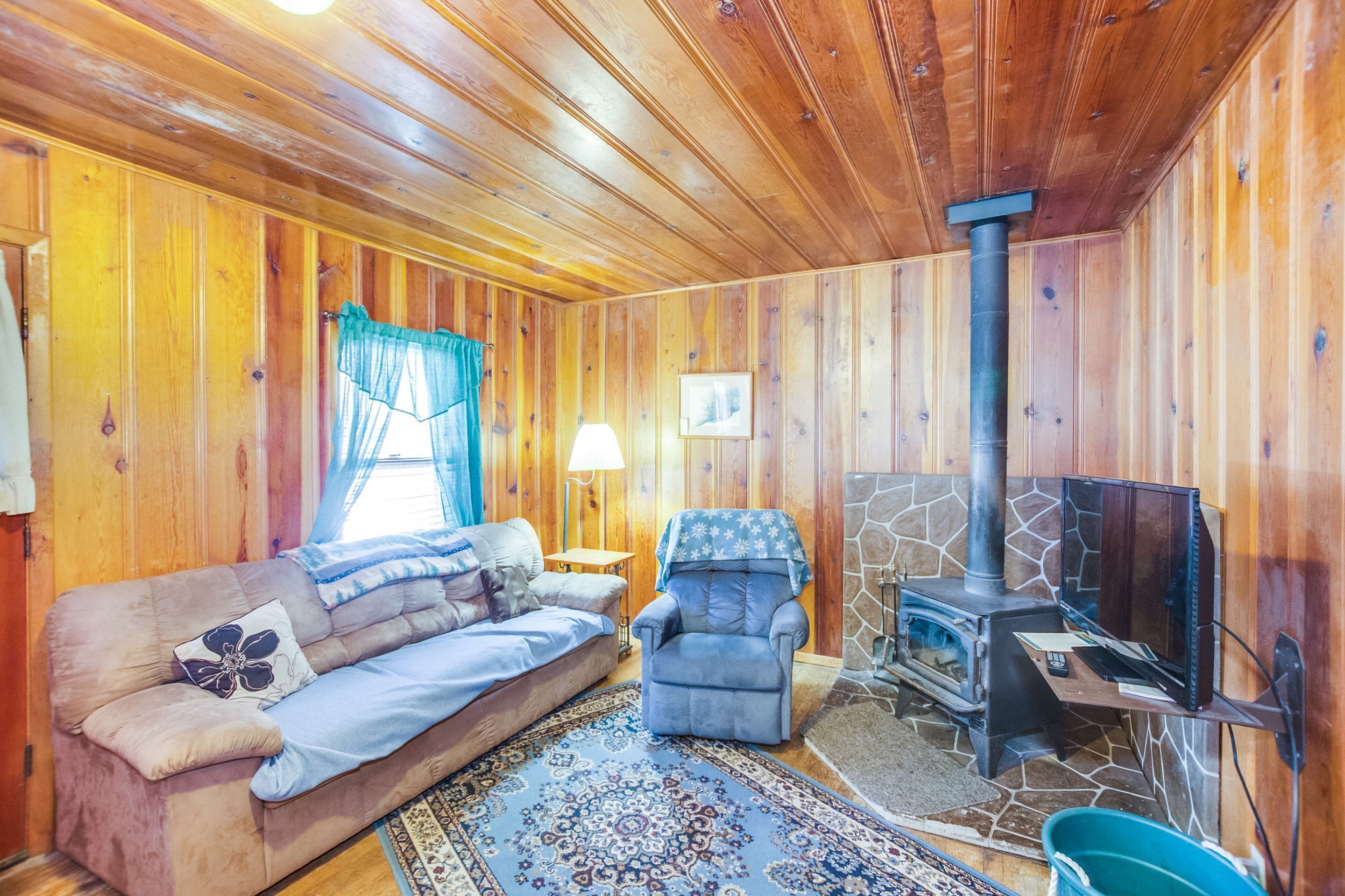 Interior acolhedor de uma cabana, com lareira de pedra, sofá, poltrona e televisão.