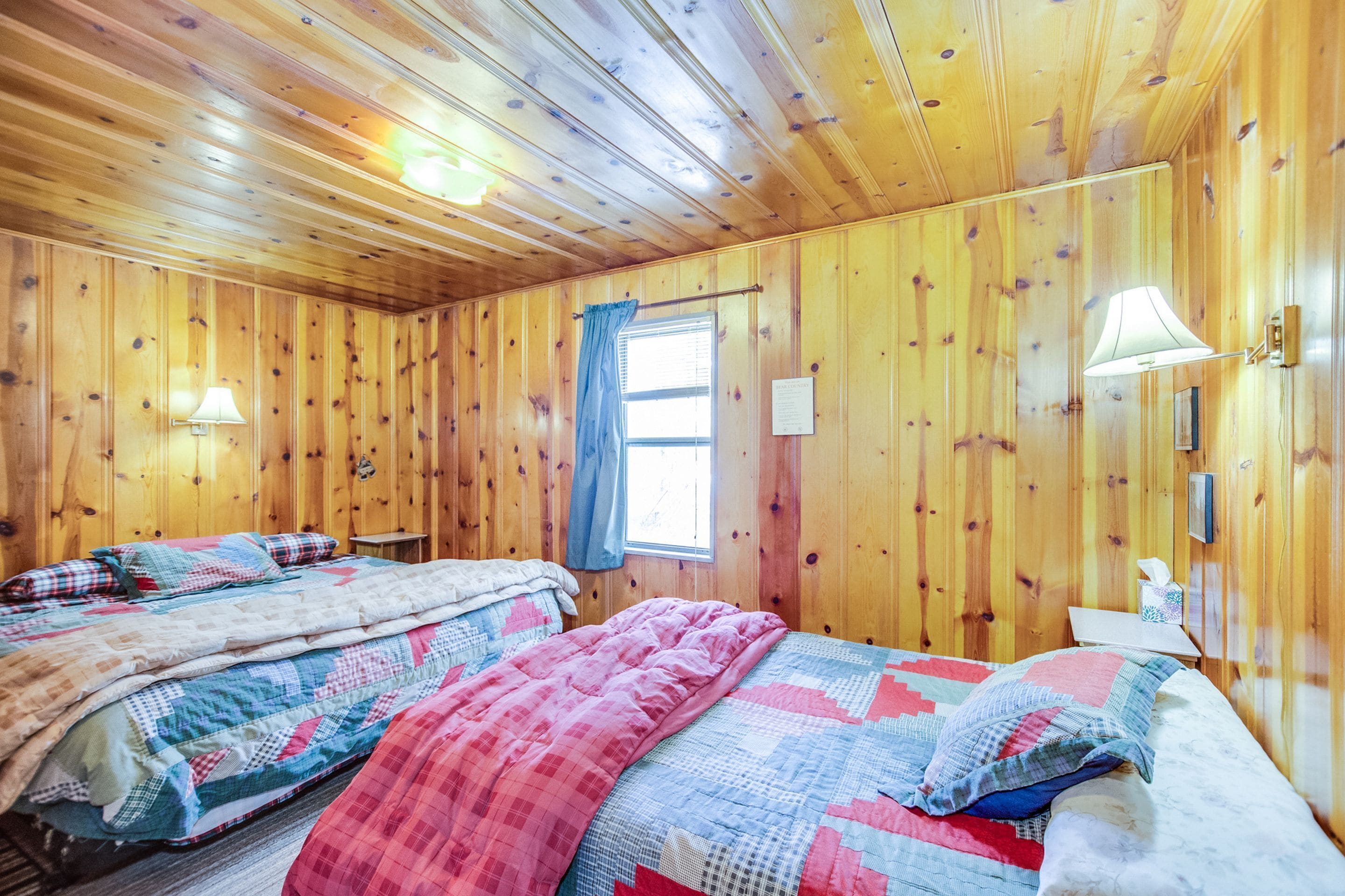 A wooden cabin room with two beds, a window with a curtain, and a nightstand with a lamp.