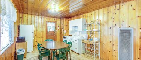 A compact kitchen with wooden walls, a dining table with chairs, a refrigerator, a stove, and a coffee maker.