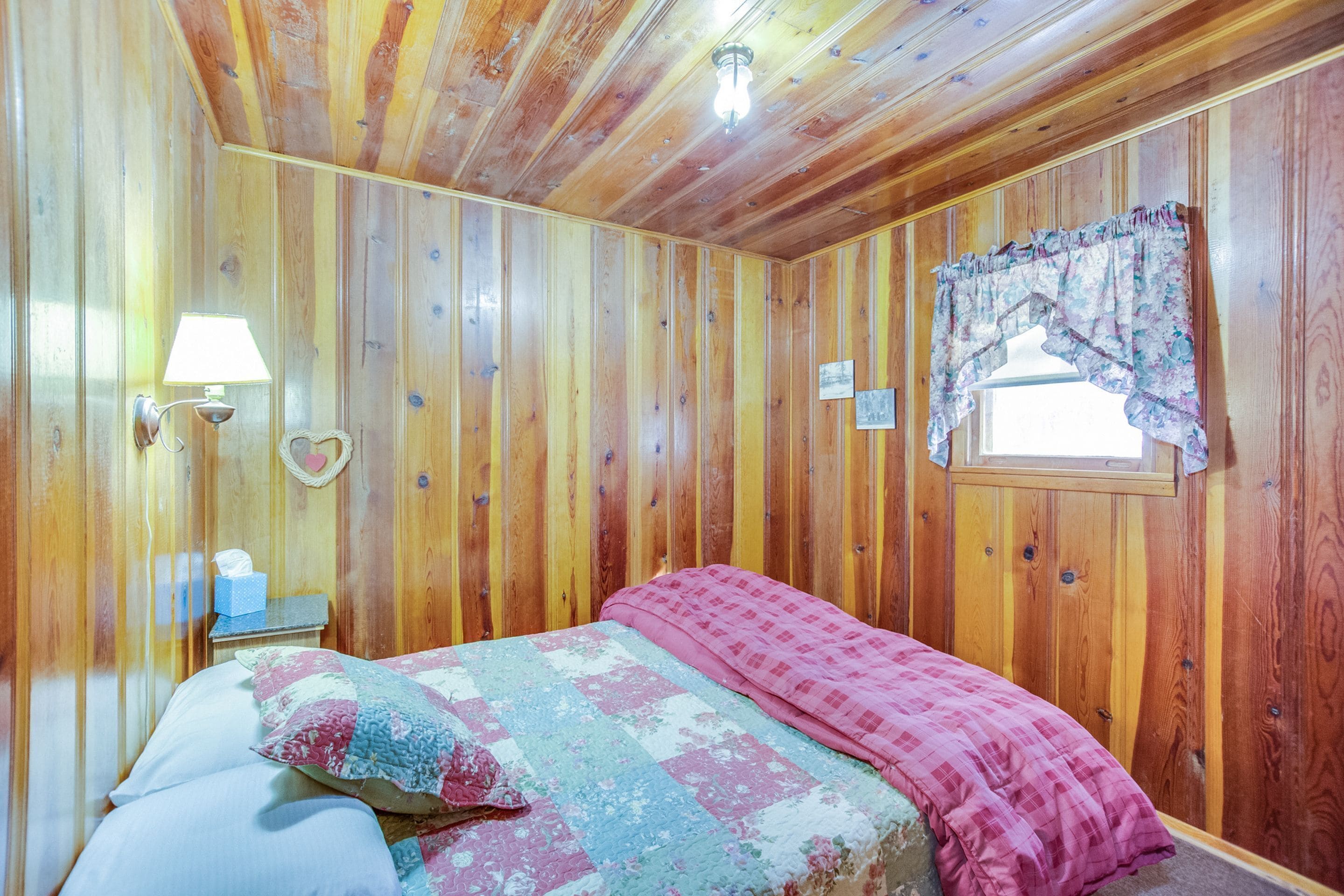 A wooden-paneled room with a bed, a nightstand, a lamp, and a window with a curtain.