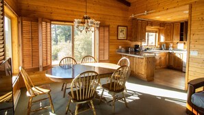 Interior de una cabaña de madera con una mesa de comedor, sillas, un candelabro, una zona de cocina y ventanas amplias.