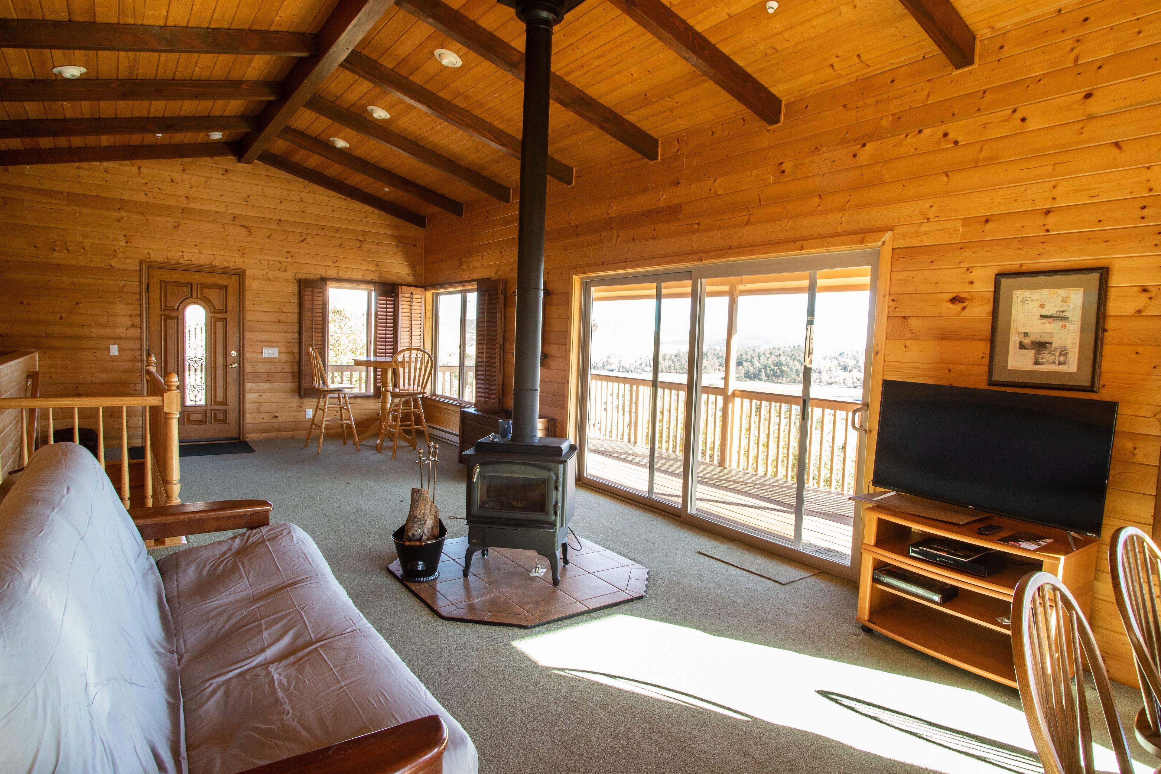 A wooden cabin interior with a sofa, a television, a fireplace, and large windows.