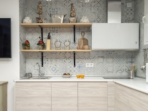 Cozinha com bancada branca, armários de madeira, pia de aço inoxidável e objetos decorativos nas prateleiras.