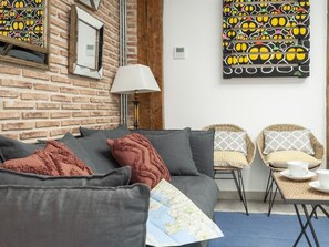 Une pièce chaleureuse avec un mur en briques, un canapé avec des coussins, deux fauteuils en osier, une petite table avec des tasses et une lampe fixée au mur.