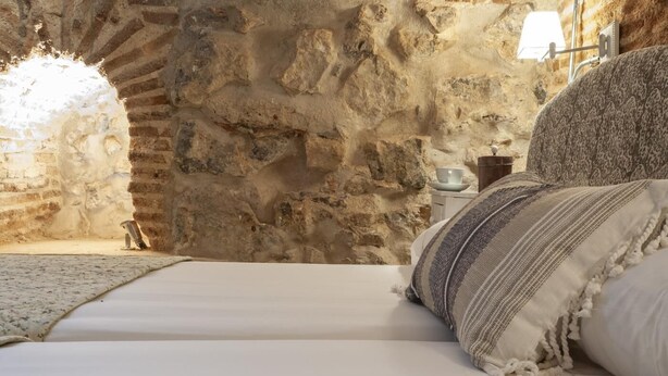 A bedroom with a stone wall, a bed with white linens, a patterned pillow, and a small bedside table with a cup and saucer. - Fantastic apartment in Barrio de las Letras (Madrid)