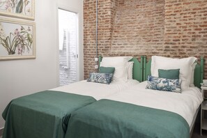 Una cama doble con ropa de cama verde, una pared de ladrillo e impresiones botánicas enmarcadas en la pared.