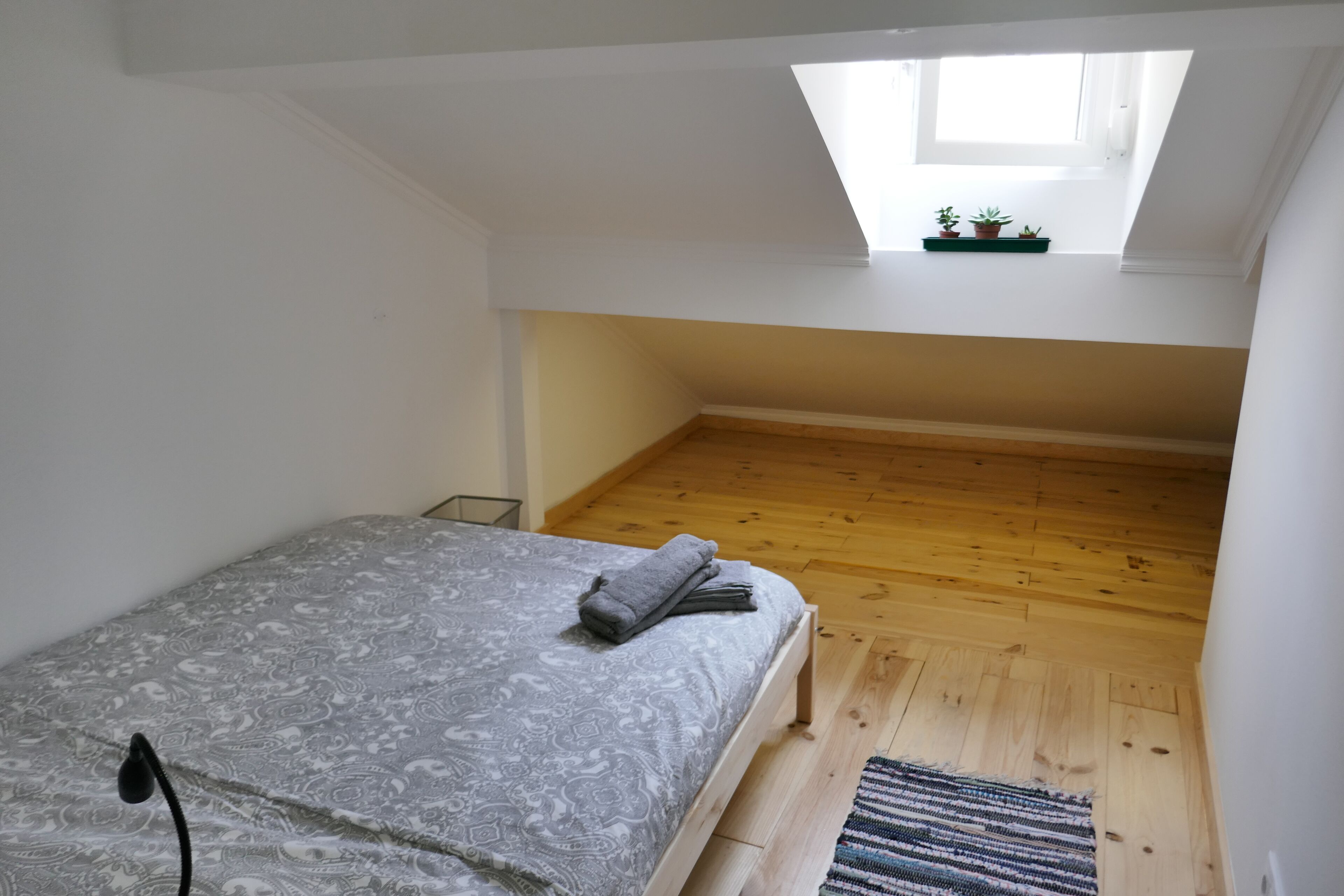 Quarto com teto inclinado, claraboia, cama com colcha estampada e pequena janela com plantas.