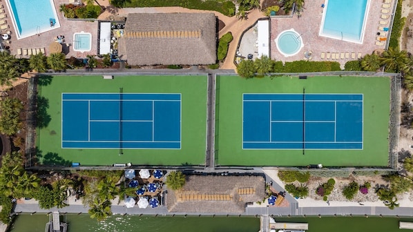 Vue aérienne d’un complexe hôtelier comprenant deux courts de tennis, un espace piscine et une plage.