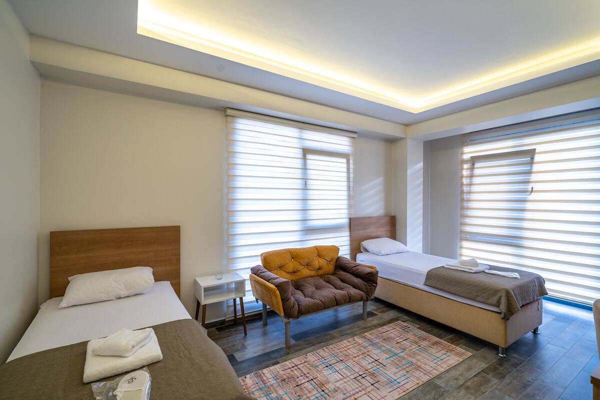 A modern hotel room with a large bed, a smaller bed, a sofa, a nightstand, and a patterned rug.