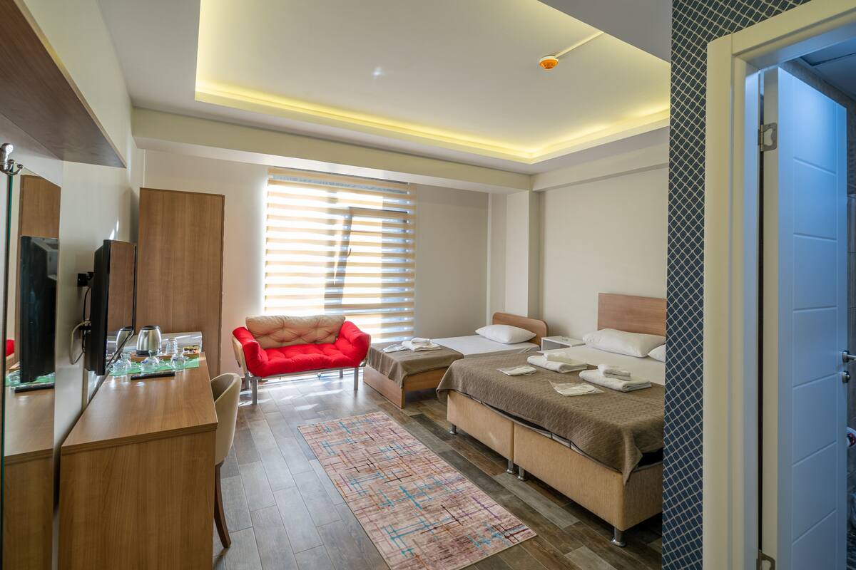 A modern hotel room with a wooden desk, a red armchair, a bed with a headboard, and a large window with blinds.