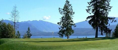 Un campo de golf con una bandera, árboles y montañas al fondo.