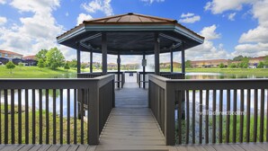 A gazebo with a wooden deck leading to it, surrounded by a lake and greenery. - Mickeys Lodge by VillaDirect (Kissimmee)