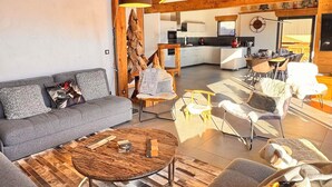 Een ruime woonkamer met een hoog houten plafond, een grijze zithoek en een ronde salontafel.