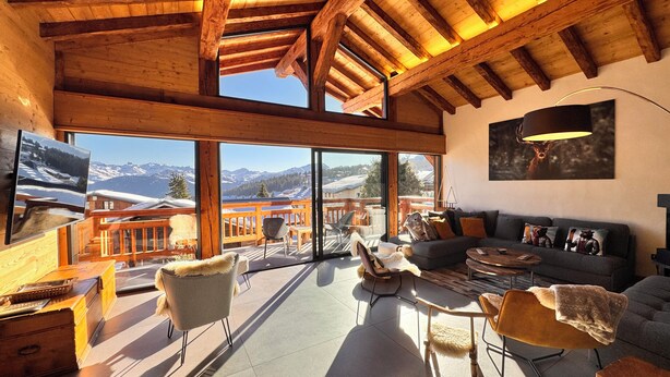 Een ruime woonkamer met een groot raam met uitzicht op een besneeuwd berglandschap. Er is een houten balkon met een leuning, en de kamer is voorzien van een bank, stoelen en een salontafel.