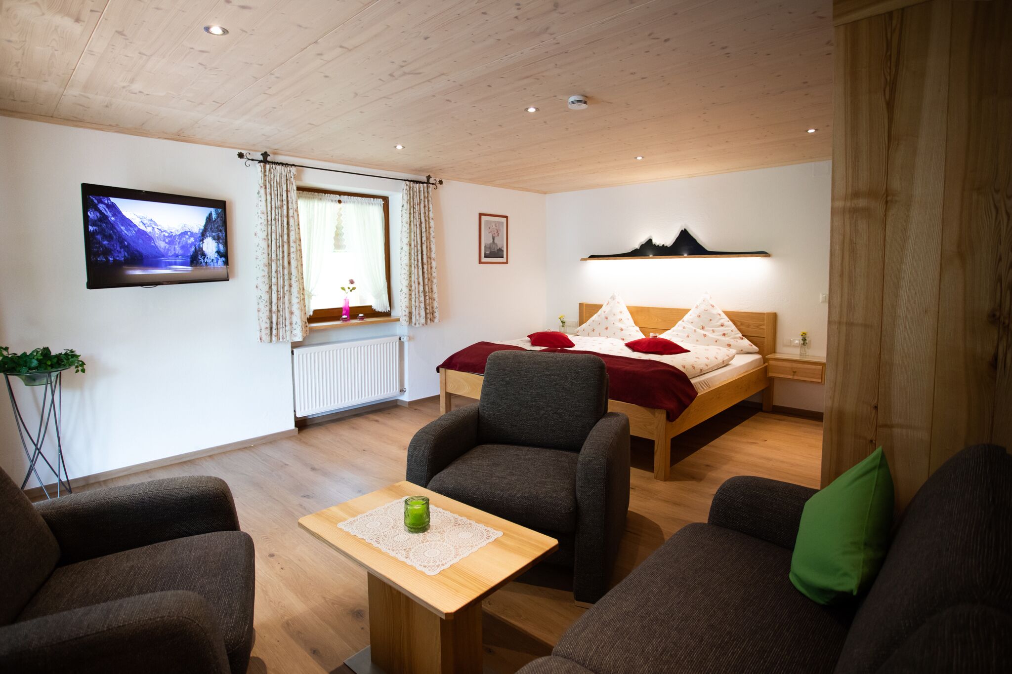 A cozy room with a bed, a TV, a window with curtains, a small table with a candle, and a sofa.