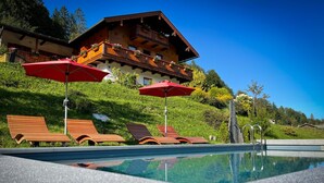 Un área de piscina con tumbonas de madera y sombrillas rojas, rodeada de exuberante vegetación y un edificio de madera con balcones.