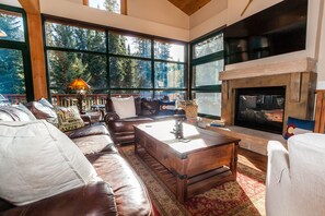 Relax in a spacious living room with a high ceiling and forest views, featuring a leather sofa set and a modern fireplace.