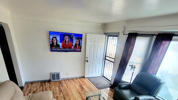Een woonkamer met een beige bank, een donkergrijze bank en een glazen salontafel. Een flatscreen televisie aan de muur toont een nieuwsuitzending.