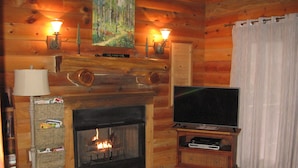 A cozy log cabin interior with a fireplace, a television on a wooden stand, and a wall-mounted lamp. - Riverfront, pet friendly ! Log cabin with 2 bedrooms and loft! (Luray)
