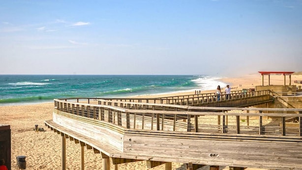 Een houten looppad dat naar een strand leidt, met een rode constructie aan de rechterkant.