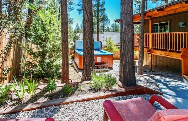 A wooden cabin with a deck, surrounded by trees and a garden area with a hot tub.