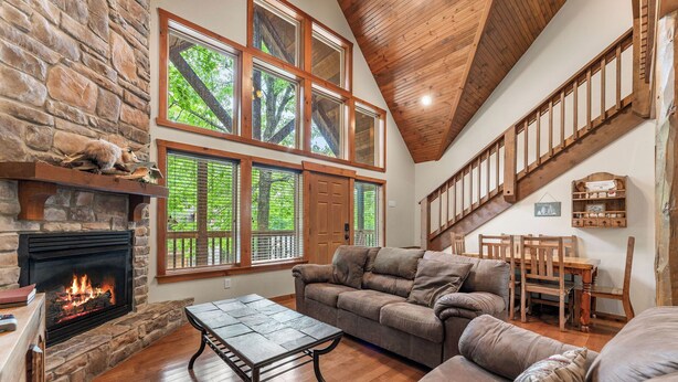 A spacious living room with a stone fireplace, large windows, a wooden ceiling, and a cozy seating area. - Close to SCD, Both Indoor & Outdoot pools, Possum Tracks Lodge  4BR/4BA Lodge (Branson West)