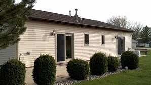 Une maison de plain-pied avec un revêtement extérieur de couleur beige, une porte d’entrée dotée d’un cadre noir et deux cheminées sur le toit. Devant la maison, on trouve quatre arbustes bien entretenus, ainsi qu’un petit sentier de gravier menant à la porte.