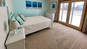 Une chambre à coucher comprenant un lit, une table de chevet, une commode, un ventilateur de plafond et une porte-fenêtre coulissante donnant sur un balcon.