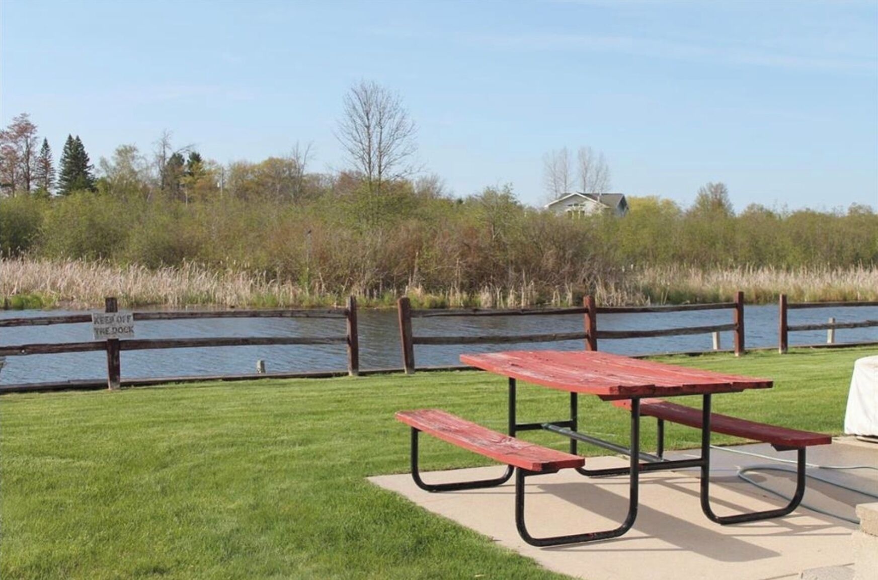 A picnic table with benches in a grassy area near a body of water, with a wooden fence and a sign that reads "KEEP OFF THE DOCK."