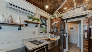 A modern kitchen with granite countertops, stainless steel appliances, and wooden cabinetry. There are decorative pineapples and plants on the shelves, and a 'relax' sign above the sink. - Newly Built Tiny House Near College Station and Round Top Area (Caldwell)